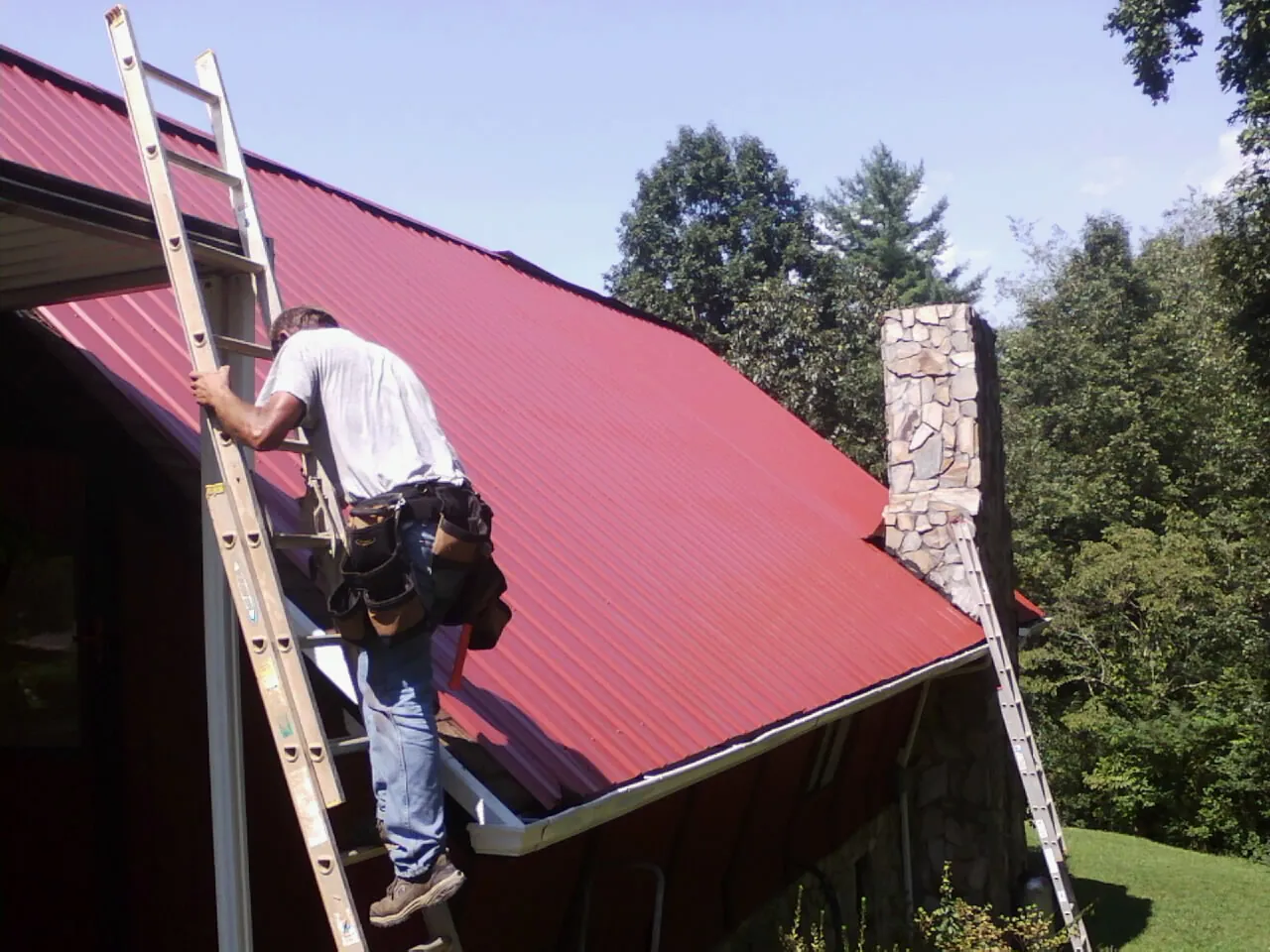 Roofer on a ladder beside a red metal roof and stone chimney — Soffit & Fascia Repair by Roofing Brothers in Rio Rancho